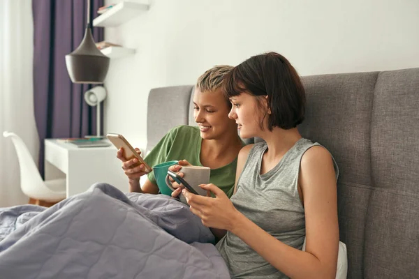 Lesbianas usan teléfonos inteligentes y beben café en la cama —  Fotos de Stock