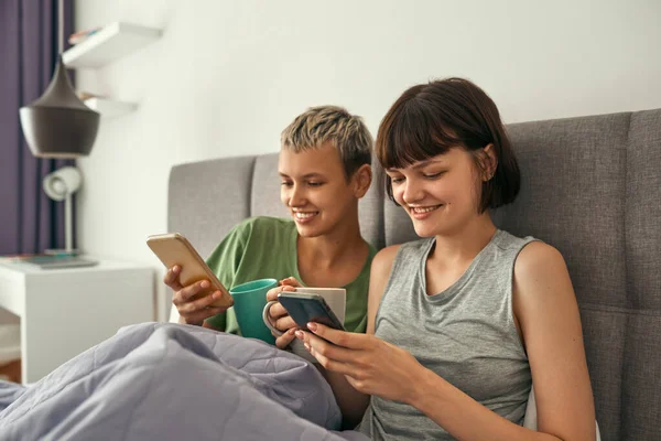 Lesbianas con té viendo en los teléfonos móviles en la cama —  Fotos de Stock
