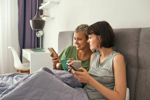 Lesbianas con té usando smartphones en la cama en casa —  Fotos de Stock