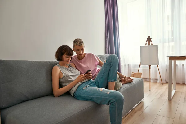 Lesbian girls watch on smartphone on sofa at home — Fotografia de Stock