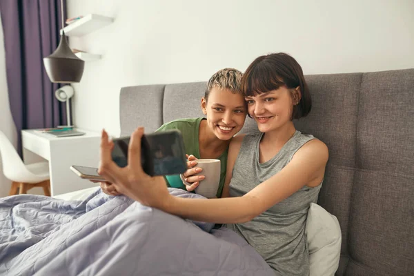Lesbians with tea take selfie on smartphone on bed — Fotografia de Stock