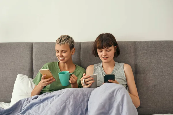 Lesbianas usando smartphones y tomando té en la cama —  Fotos de Stock