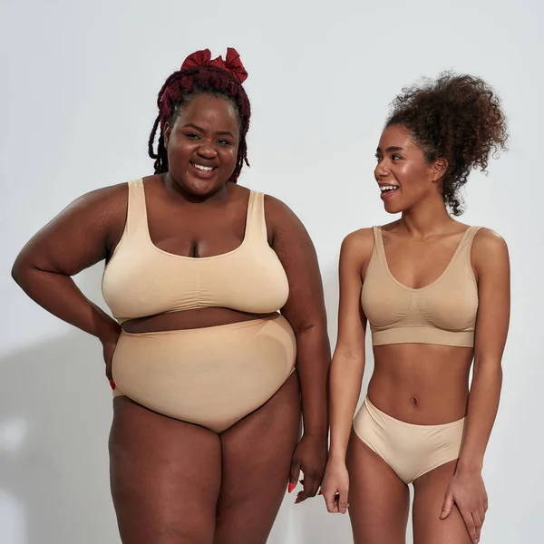 Duas mulheres americanas africanas magras e gordas confiantes em roupa interior bege sorrindo enquanto estão juntas isoladas sobre fundo cinza — Fotografia de Stock