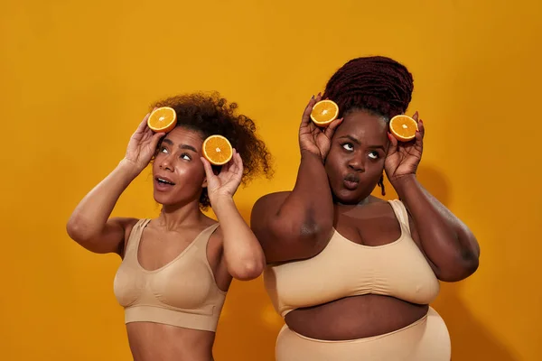 Dos mujeres americanas africanas juguetonas luciendo tontas, sosteniendo la mitad de naranja madura, posando juntas aisladas sobre fondo naranja — Foto de Stock