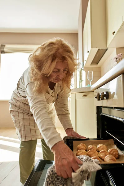 年配の女性はオーブンからクロワッサンとトレイを取ります — ストック写真