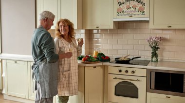 Gri saçlı çift yemek yaparken beyaz şarap içiyor.