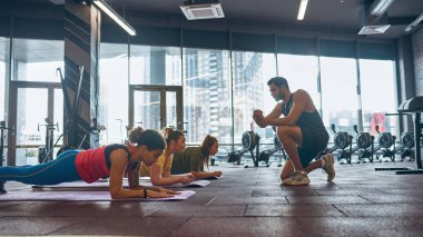 Eğitmen kalas egzersizi yapan kadınlara bakıyor.