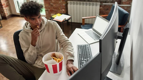 Afro-americano joven trabajador independiente comiendo papas fritas en el almuerzo mientras usa pc —  Fotos de Stock