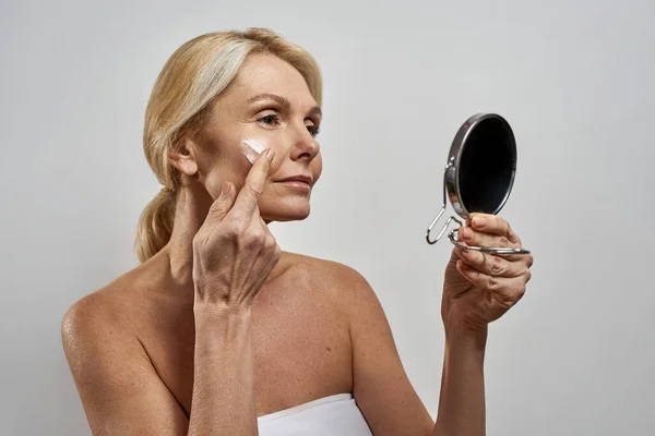 Mulher olha para o espelho enquanto esfregaço creme no rosto — Fotografia de Stock