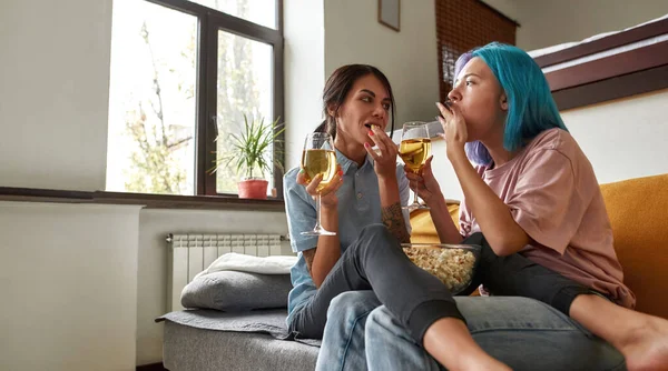 Duas meninas comendo pipocas e bebendo vinho no sofá — Fotografia de Stock