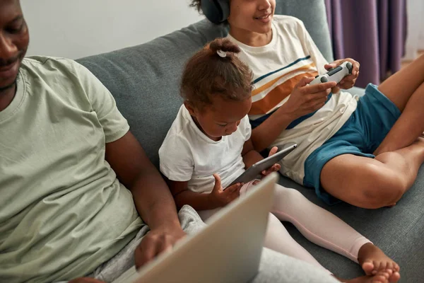 Preto pai e crianças usam gargets no sofá em casa — Fotografia de Stock