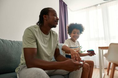 Çocuk video oyununda babasının kumandasındaki düğmeye basıyor.