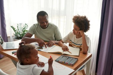 Babası oğluna ödevlerinde yardım ederken kızı resim çiziyor.