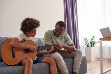 Baba, oğluna akustik gitar çalmayı öğretiyor ve yardım ediyor.