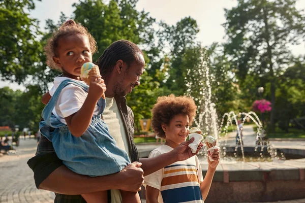 Černá rodina jíst zmrzliny v blízkosti fontány v parku — Stock fotografie