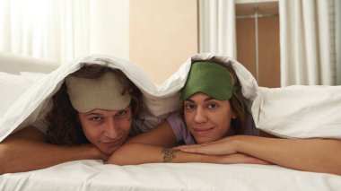 Yatakta uzanmış kameraya bakan gülümseyen bir çift.