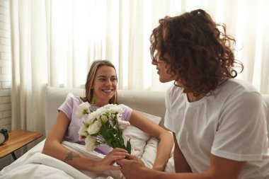 Yatakta gülümseyen kız arkadaşına çiçek veren adam.