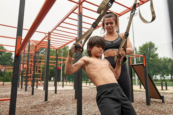 Kadın, oğluna süspansiyon kayışlarıyla egzersiz yapmayı öğretiyor.