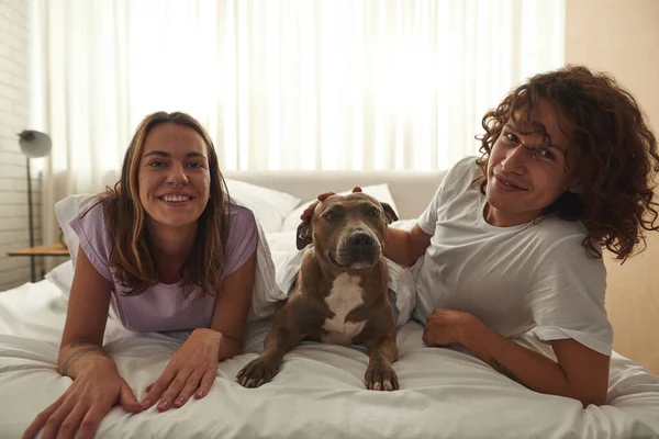Joven sonriente pareja caucásica y perro yacía en la cama — Foto de Stock