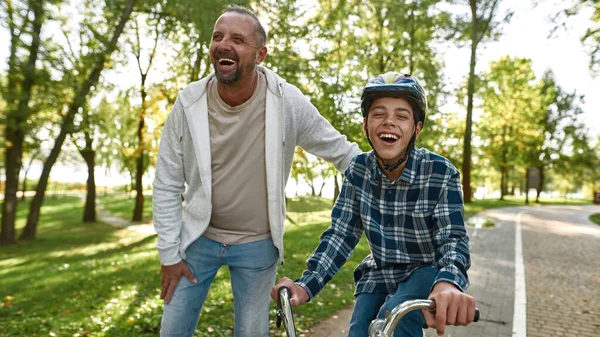 Father helping son riding bicycle in sunny park — Zdjęcie stockowe