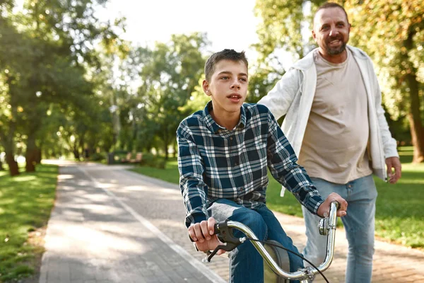 Father helping teenage son riding bicycle in park — Foto de Stock