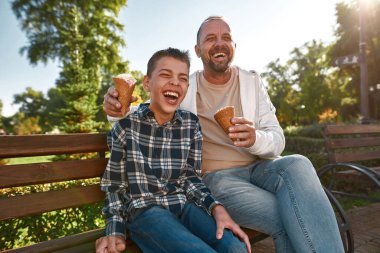 Neşeli baba ve oğul güneşli parkta dondurma yiyorlar.