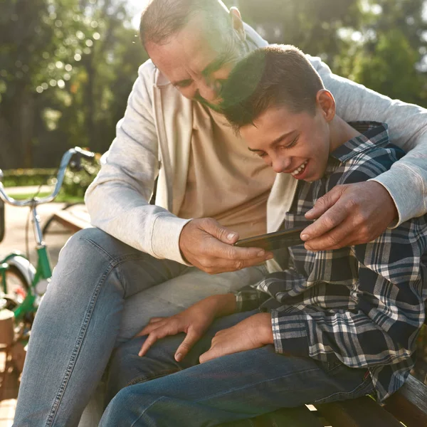 Father and son watch smartphone on bench in park — Stok fotoğraf