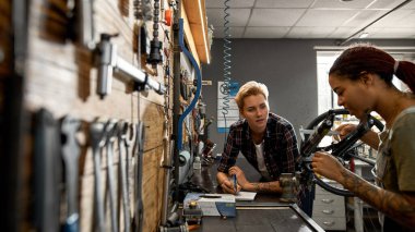 Not defterine notlar yazan bir kız ve atölyedeki kalibreli bisiklet çatalını ölçen meslektaşına bakıyor. Çok ırklı genç kadın zanaatçılar. Takım çalışması. Bisiklet servisi, tamir ve geliştirme