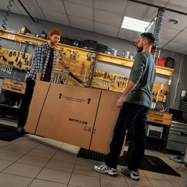 Erkek işçiler atölyede karton kutu ve yeni bisiklet taşıyorlar. Genç, beyaz bisikletli tamirciler. Takım çalışması. Bisiklet servisi, tamir ve geliştirme. Araç gereç ve ekipman ile garaj içi