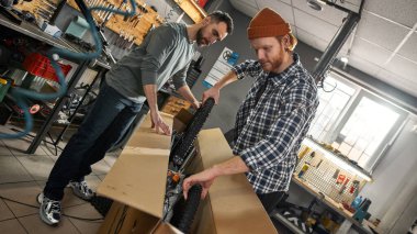 Atölyedeki karton kutudan yeni bir bisikletin tekerleklerini alan adamlar. Genç Kafkas bisiklet ustaları. Takım çalışması. Bisiklet servisi, tamir ve geliştirme. Araç gereç ve ekipman ile garaj içi