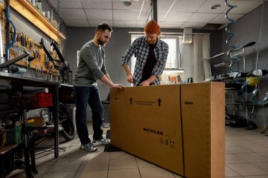 Erkek işçiler atölyede karton kutuyu yeni bisikletle açıyorlar. Genç beyaz bisiklet teknisyenleri. Takım çalışması. Bisiklet servisi, tamir ve geliştirme. Araç gereç ve ekipman ile garaj içi
