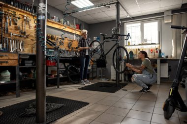 Two female cycling mechanics fixing or installing wheels on bicycle in workshop. Young multiethnic women colleaugues. Bike service, repair and upgrade. Garage interior with tools and equipment clipart