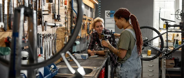 Woman writing notes in notebook about his colleague measuring bicycle fork with calipers in workshop. Young multiracial female servicemen. Teamwork. Bike service, repair and upgrade