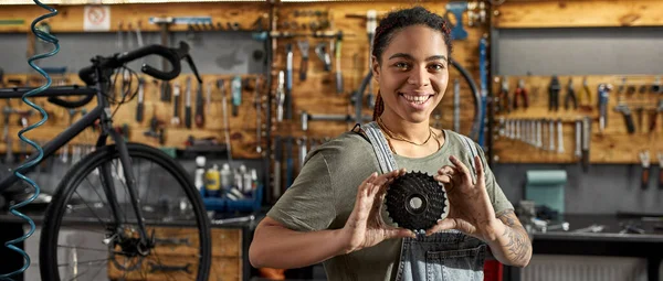 汚い手で保持し ワークショップで自転車カセットを示す女性サイクリング技術者 若い黒人の笑顔の女性がカメラを見ている 自転車サービス 修理とアップグレード 工具付きガレージインテリア — ストック写真