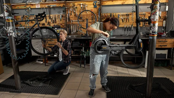 Black woman mechanic fixing electric scooter with screwdriver near female caucasian colleague installing wheel of bicycle with wrench in modern workshop. Bike service, repair and upgrade