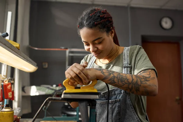 Female cycling master fixing bike tube in bicycle workshop. Young smiling african american girl with tattoos. Bike service, repair and upgrade. Garage interior with tools and equipment