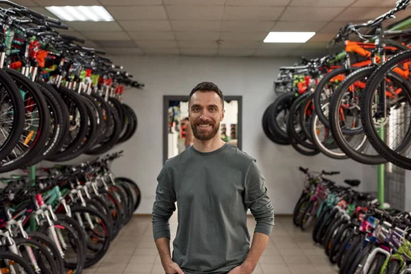 Front view of young smiling caucasian bearded male seller with hands in pockets looking at camera in modern bike shop. Set of variety bikes for extreme sport, urban lifestyle and children