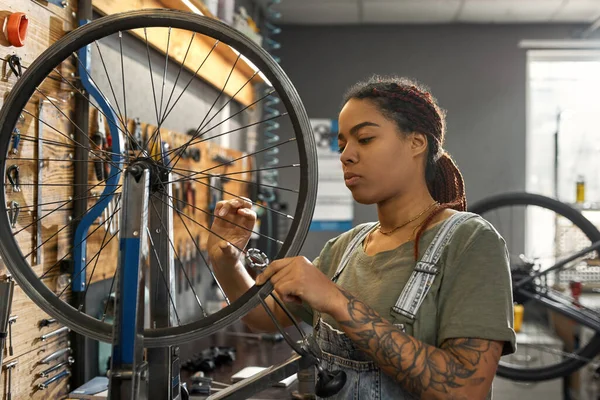 若いアフリカ系アメリカ人女性の自転車整備士のチェック自転車ホイールは 現代のワークショップで重要な話をした 自転車サービス 修理とアップグレード 設備付きガレージインテリア — ストック写真