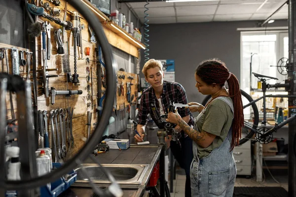 Woman writing notes in notebook about his colleague measuring bicycle fork with calipers in workshop. Young multiracial female cycling repairmen. Teamwork. Bike service, repair and upgrade