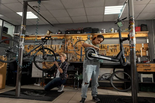 Black woman repairman fixing electric scooter with screwdriver near female caucasian colleague installing wheel of bicycle with wrench in modern workshop. Bike service, repair and upgrade