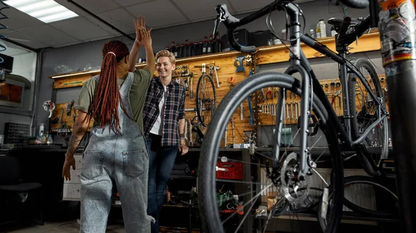 自転車のワークショップでは2人の女性修理生が互いに5人ずつ与え合う 若い多民族女性の同僚 自転車サービス 修理とアップグレード 工具や機器とガレージのインテリア 友情だ チームワーク — ストック写真