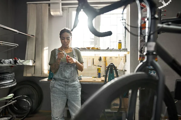 Female cycling mechanic cutting patch with scissors for glueing on bike tube in modern bicycle workshop. Young concentrated black girl. Bike service, repair and upgrade. Sunny daytime
