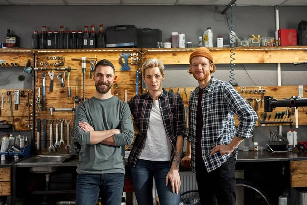 Team of three cycling workers posing and looking at camera at workplace in modern bicycle workshop. Young caucasian men and woman. Friendship. Teamwork. Bike service, repair and upgrade