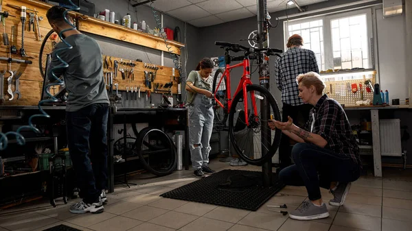 Team of four multiracial cycling workers working in modern bicycle workshop. Young men and woman. Teamwork. Bike service, repair and upgrade. Garage interior with tools and equipment