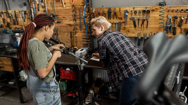 Woman writing notes in notebook and looking at his colleague measuring bicycle fork with calipers in workshop. Young multiracial female servicemen. Teamwork. Bike service, repair and upgrade