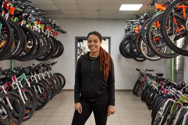 Front view of young smiling african american female seller posing and looking at camera in modern bike shop. Set of variety bikes for extreme sport, urban lifestyle and children. Shopping concept