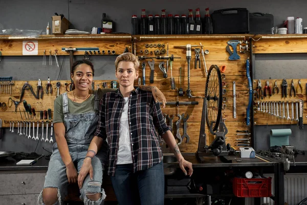 ワークショップにポーズをとる2人の若い多民族自転車力学 女性労働者の同僚抱擁とカメラを見て 自転車サービス 修理とアップグレード 工具や機器とガレージのインテリア — ストック写真