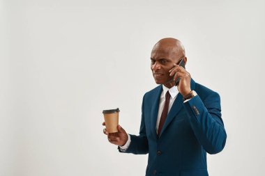 Elinde kahve tutan ve akıllı telefondan konuşan şüpheci yüzlü Afro-Amerikalı iş adamı. Takım elbiseli kel bir yetişkin. Modern başarılı erkek yaşam tarzı. Beyaz arka plan. Stüdyo çekimi. Boşluğu kopyala