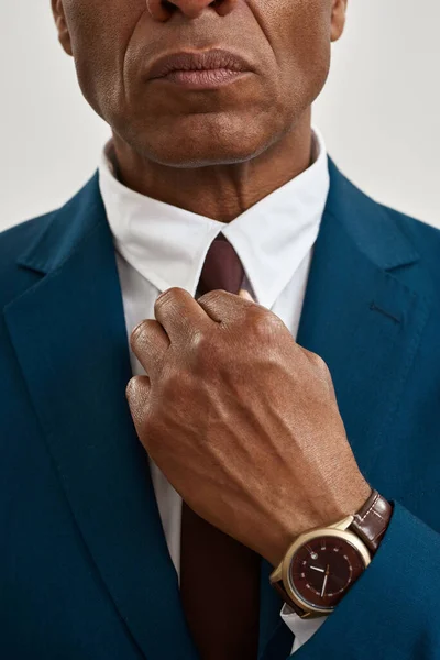 Cropped image and obscure face of serious adult black businessman straighten tie of his formal wear. Concept of modern successful male lifestyle. Isolated on white background. Studio shoot