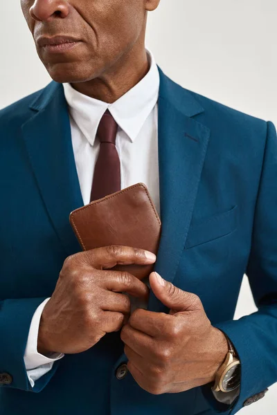 Obscure face of serious adult black entrepreneur or company leader putting wallet inside suit pocket. Concept of modern successful male lifestyle. Isolated on white background. Studio shoot